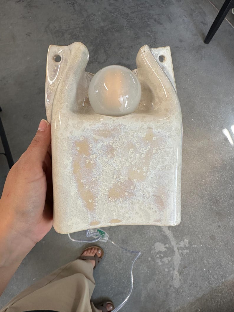 May include: A wall-mounted ceramic sconce with a glossy, cream-coloured speckled glaze. The fixture features a U-shaped cutout at the top, two circular mounting holes, and a single round, glowing light bulb centred within the ceramic housing.