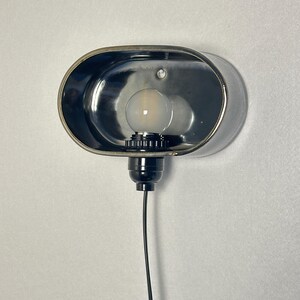 K&ouml;nnte beinhalten: Eine verchromte Wandleuchte mit ovalem Schirm. Die Leuchte hat einen schwarzen Sockel, eine Gl&uuml;hbirne und ein schwarzes Elektrokabel. An einer wei&szlig;en Wand montiert, bietet sie eine moderne Beleuchtungsl&ouml;sung.