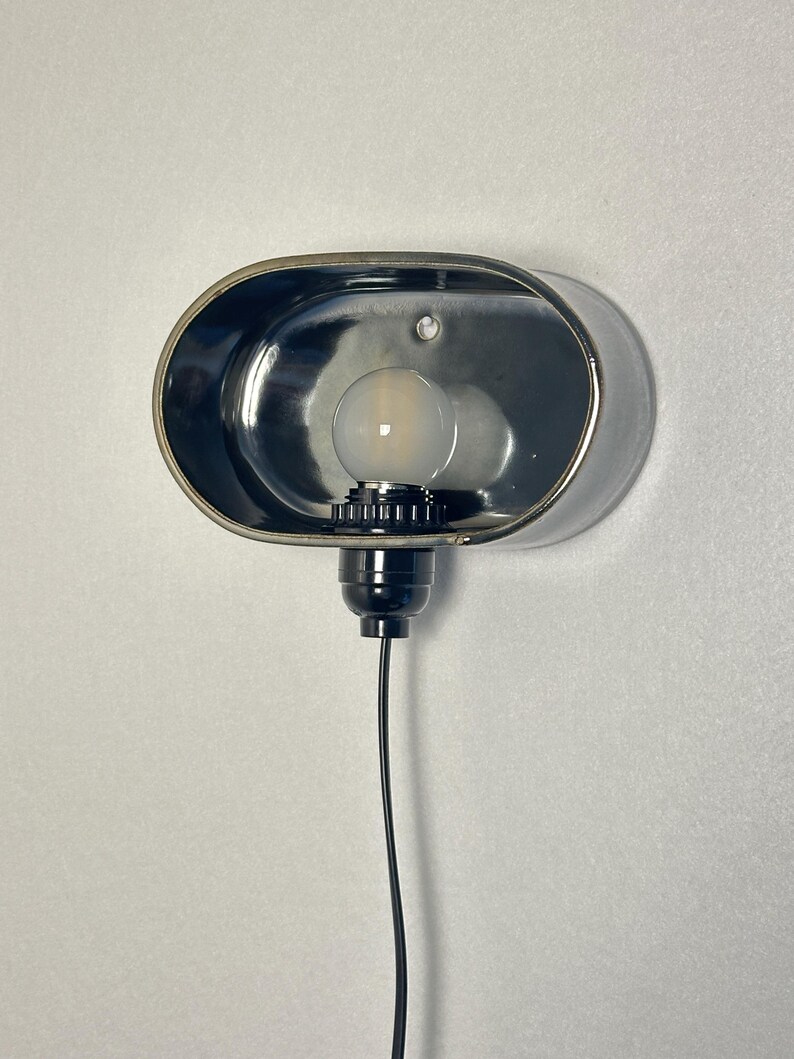 K&ouml;nnte beinhalten: Eine verchromte Wandleuchte mit ovalem Schirm. Die Leuchte hat einen schwarzen Sockel, eine Gl&uuml;hbirne und ein schwarzes Elektrokabel. An einer wei&szlig;en Wand montiert, bietet sie eine moderne Beleuchtungsl&ouml;sung.