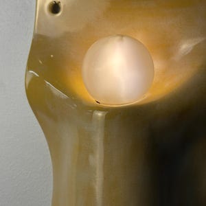 May include: A wall-mounted ceramic sconce with a glossy, mustard-yellow glaze and an organic, curved silhouette. The fixture features a single circular opening at the top for mounting and a central, glowing spherical light bulb.