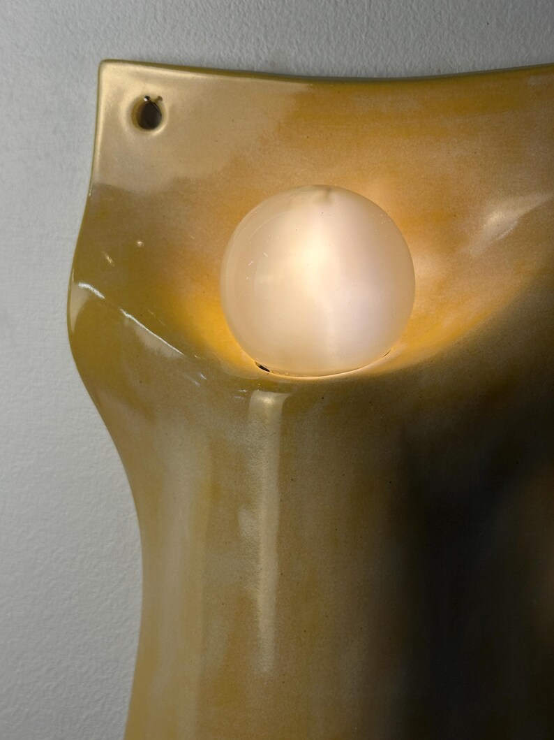 May include: A wall-mounted ceramic sconce with a glossy, mustard-yellow glaze and an organic, curved silhouette. The fixture features a single circular opening at the top for mounting and a central, glowing spherical light bulb.