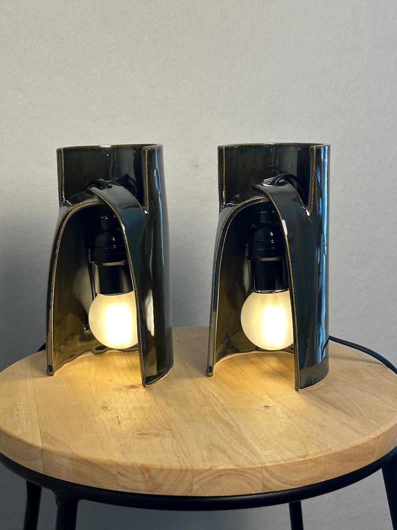 Puede incluir: Un par de l&aacute;mparas de mesa de cer&aacute;mica oscura y brillante con una silueta arquitect&oacute;nica curva. Cada l&aacute;mpara cuenta con un casquillo negro que sostiene una bombilla redonda esmerilada, creando una iluminaci&oacute;n c&aacute;lida en su interior.
