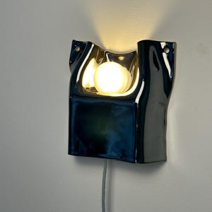 May include: A wall-mounted lamp featuring a glossy, dark blue ceramic shade with an irregular, folded shape. A single, round, warm-toned light bulb is positioned in the center of the shade, emitting a soft glow. A thin power cord hangs below.