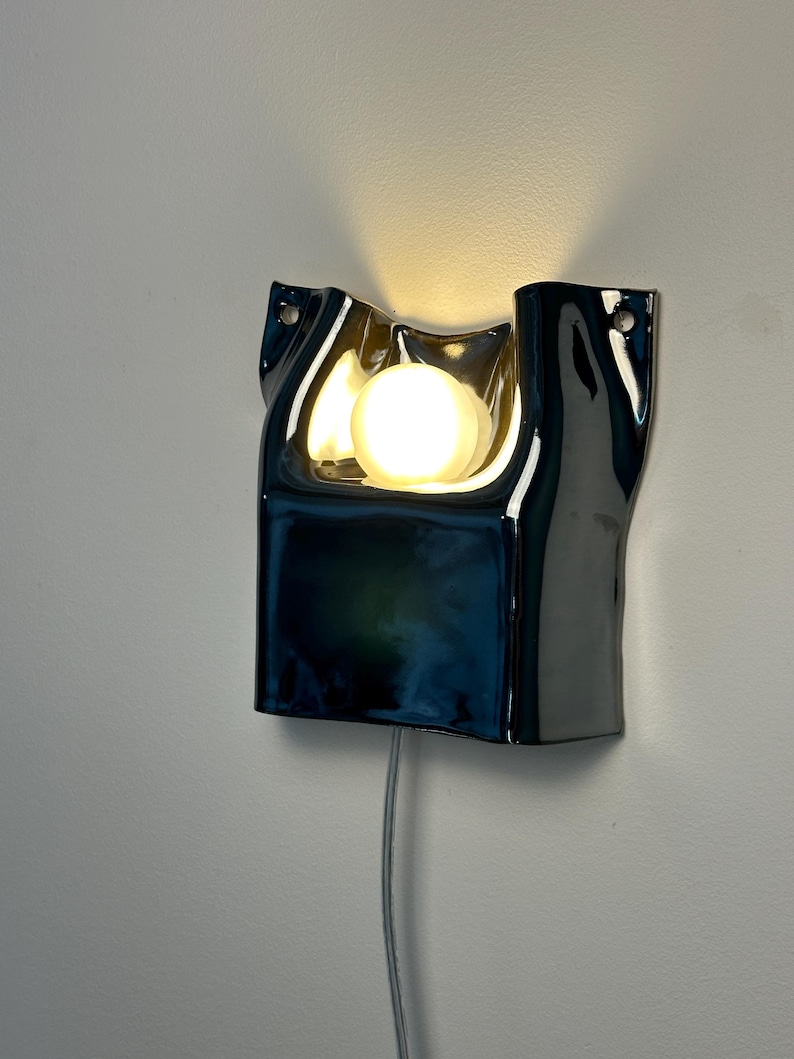 May include: A wall-mounted lamp featuring a glossy, dark blue ceramic shade with an irregular, folded shape. A single, round, warm-toned light bulb is positioned in the center of the shade, emitting a soft glow. A thin power cord hangs below.