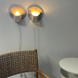 May include: Two wall-mounted sconces with a speckled grey ceramic finish and a shell-like shape. Each light fixture emits a warm, glowing light from the centre and includes a thin white power cord hanging down towards the floor.