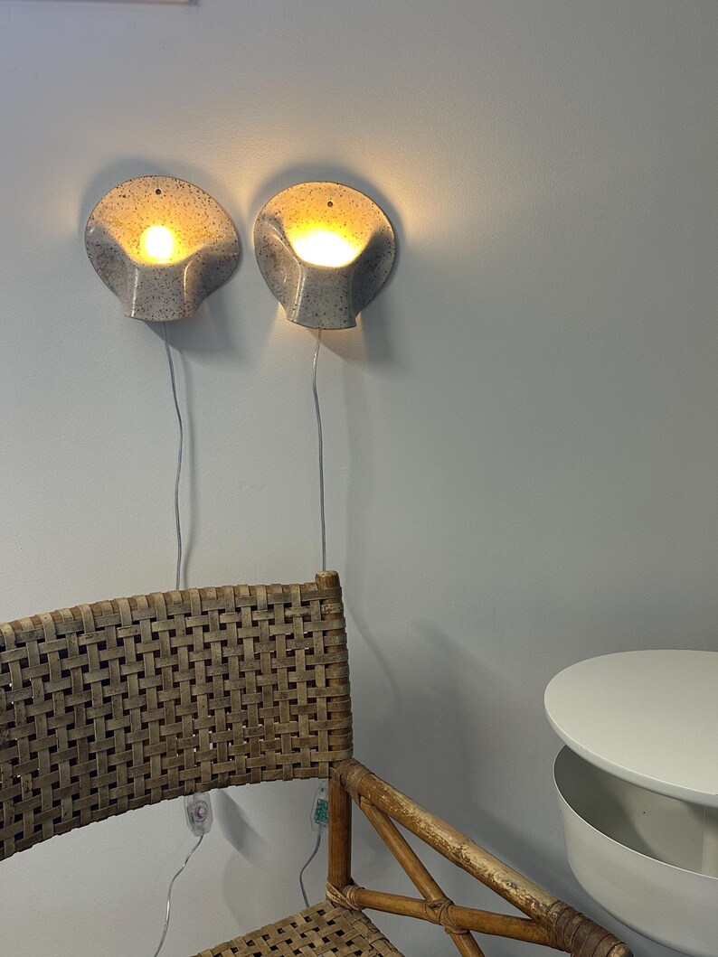 May include: Two wall-mounted sconces with a speckled grey ceramic finish and a shell-like shape. Each light fixture emits a warm, glowing light from the centre and includes a thin white power cord hanging down towards the floor.