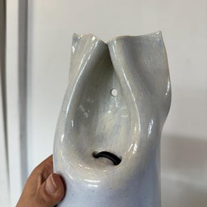 May include: A light blue ceramic wall vase with a glossy finish and an organic, folded shape. The piece features a small circular hole near the top for mounting and a larger opening below containing a black rubber ring insert for holding stems.