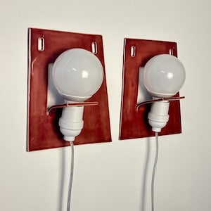 Puede incluir: Un par de apliques de pared con placas traseras rectangulares de cer&aacute;mica de color rojo oscuro brillante con esquinas redondeadas. Cada aplique incluye un casquillo blanco, una bombilla redonda blanca y un cable el&eacute;ctrico fino y claro.