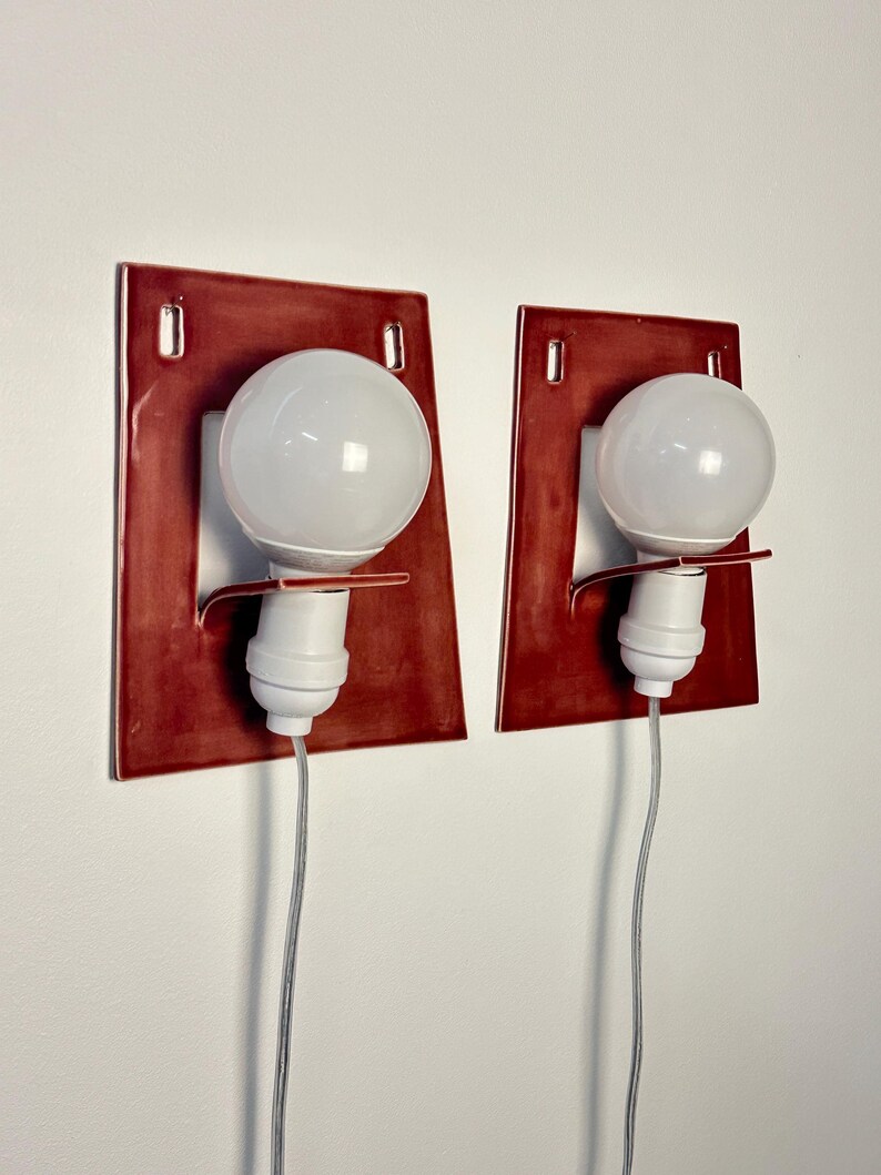 Puede incluir: Un par de apliques de pared con placas traseras rectangulares de cer&aacute;mica de color rojo oscuro brillante con esquinas redondeadas. Cada aplique incluye un casquillo blanco, una bombilla redonda blanca y un cable el&eacute;ctrico fino y claro.