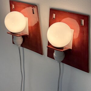 Puede incluir: Un par de apliques de pared con placas traseras cuadradas de cer&aacute;mica en color rojo oscuro brillante, con dos aberturas rectangulares en la parte superior. Cada l&aacute;mpara incluye un casquillo blanco, un peque&ntilde;o estante y una bombilla redonda.