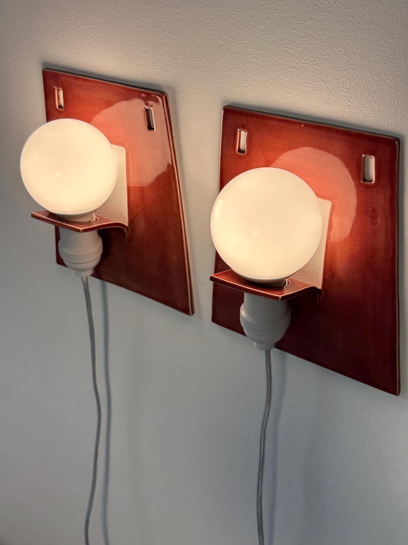 Puede incluir: Un par de apliques de pared con placas traseras cuadradas de cer&aacute;mica en color rojo oscuro brillante, con dos aberturas rectangulares en la parte superior. Cada l&aacute;mpara incluye un casquillo blanco, un peque&ntilde;o estante y una bombilla redonda.