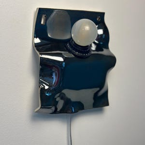 May include: A wall-mounted lamp featuring a glossy, dark, reflective, and irregularly shaped ceramic base with a rippled surface. A single, round, frosted white light bulb is centered on the front, attached to a black circular socket fixture.