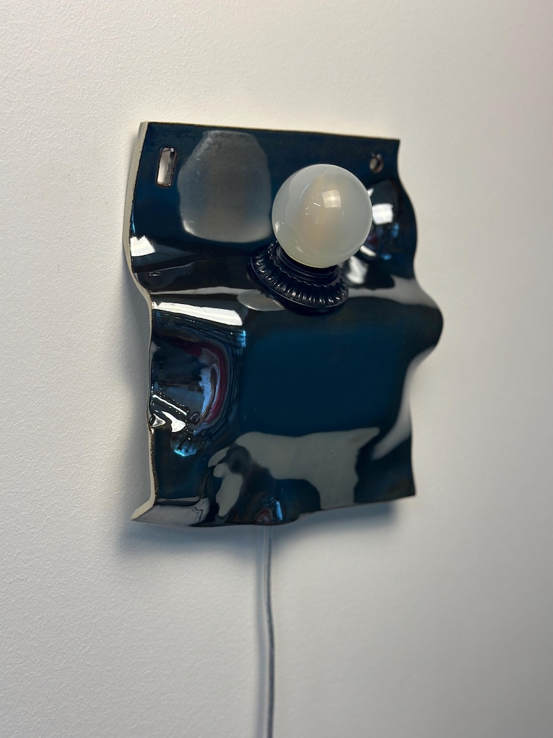May include: A wall-mounted lamp featuring a glossy, dark, reflective, and irregularly shaped ceramic base with a rippled surface. A single, round, frosted white light bulb is centered on the front, attached to a black circular socket fixture.