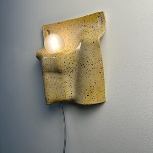 May include: A wall-mounted light fixture featuring a curved, yellow ceramic body with a black speckled pattern. The fixture has a smooth, undulating shape with two top mounting holes and a single glowing white spherical bulb nestled in a recess.