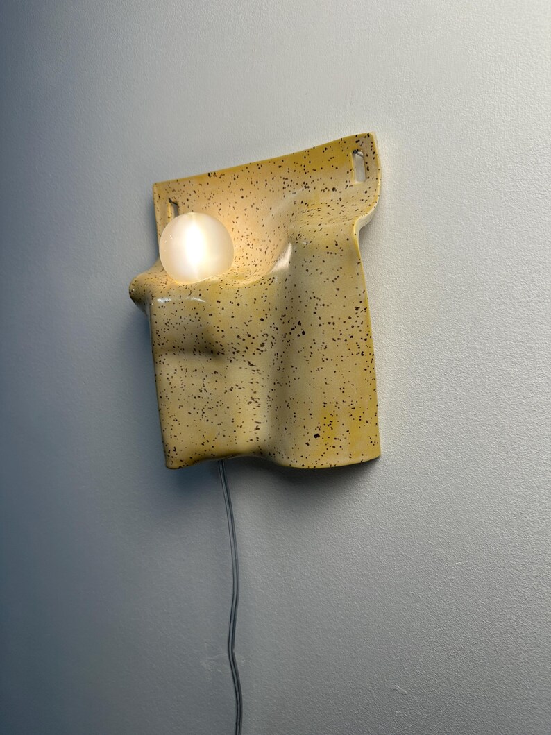 May include: A wall-mounted light fixture featuring a curved, yellow ceramic body with a black speckled pattern. The fixture has a smooth, undulating shape with two top mounting holes and a single glowing white spherical bulb nestled in a recess.