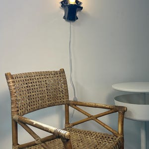May include: A woven rattan armchair with a natural light brown finish, featuring a high backrest and cross-braced armrests. Above the chair, a small black wall-mounted lamp emits a warm glow, connected by a thin white cord hanging down.