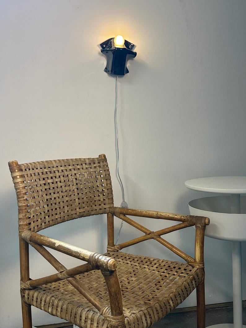 May include: A woven rattan armchair with a natural light brown finish, featuring a high backrest and cross-braced armrests. Above the chair, a small black wall-mounted lamp emits a warm glow, connected by a thin white cord hanging down.