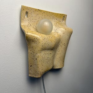 May include: A wall-mounted ceramic light fixture with a speckled yellow glaze. The piece features a curved, organic form with two rectangular mounting holes at the top and a single, central, spherical white glass light bulb extending outward.
