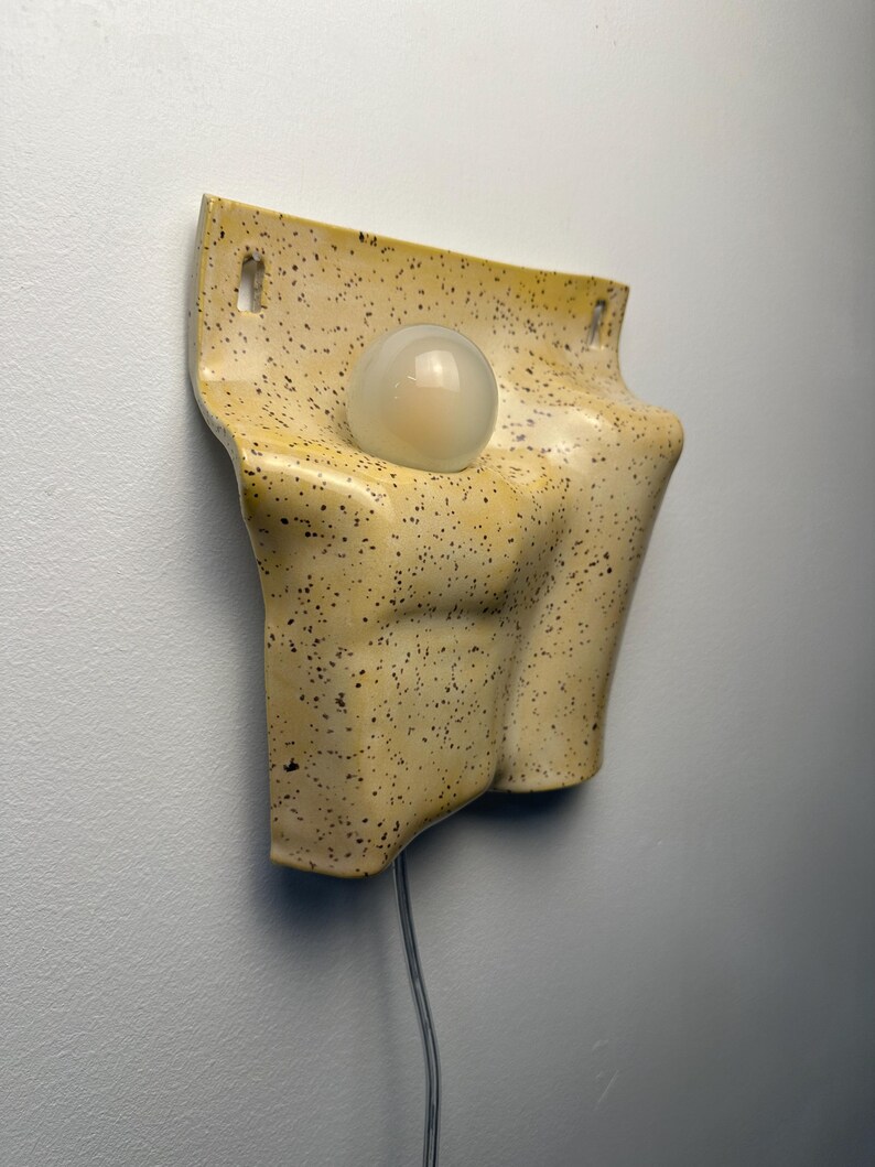 May include: A wall-mounted ceramic light fixture with a speckled yellow glaze. The piece features a curved, organic form with two rectangular mounting holes at the top and a single, central, spherical white glass light bulb extending outward.