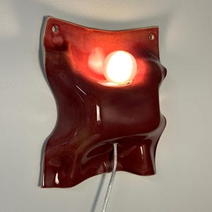 K&ouml;nnte beinhalten: Eine Wandleuchte aus Keramik mit einer gl&auml;nzenden, tiefroten, gewellten Oberfl&auml;che. Das St&uuml;ck verf&uuml;gt &uuml;ber einen zentralen kreisf&ouml;rmigen Ausschnitt mit einer leuchtenden wei&szlig;en Kugellampe, zwei kleine L&ouml;cher an den oberen Ecken und ein silbernes Kabel.