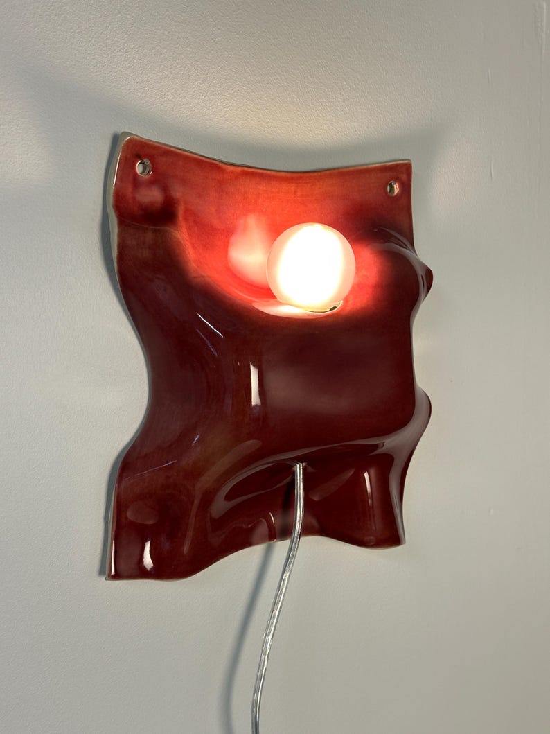 K&ouml;nnte beinhalten: Eine Wandleuchte aus Keramik mit einer gl&auml;nzenden, tiefroten, gewellten Oberfl&auml;che. Das St&uuml;ck verf&uuml;gt &uuml;ber einen zentralen kreisf&ouml;rmigen Ausschnitt mit einer leuchtenden wei&szlig;en Kugellampe, zwei kleine L&ouml;cher an den oberen Ecken und ein silbernes Kabel.