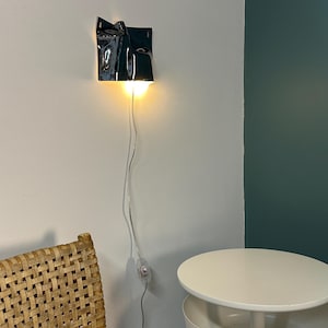 May include: A wall-mounted lamp featuring a glossy black, irregularly shaped ceramic shade with a folded, sculptural design. The lamp emits a warm, downward-facing light and includes a thin, transparent power cord with an inline switch.