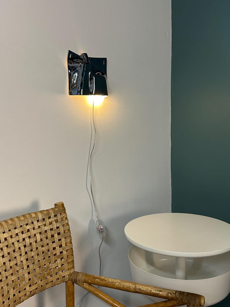May include: A wall-mounted lamp featuring a glossy black, irregularly shaped ceramic shade with a folded, sculptural design. The lamp emits a warm, downward-facing light and includes a thin, transparent power cord with an inline switch.