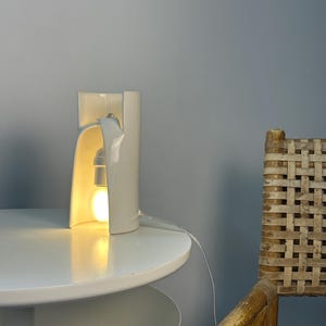 May include: A white ceramic table lamp with a curved, cylindrical structure that is open on one side. The lamp features a single light bulb emitting a warm glow from within the hollow center, connected by a thin, clear electrical cord.