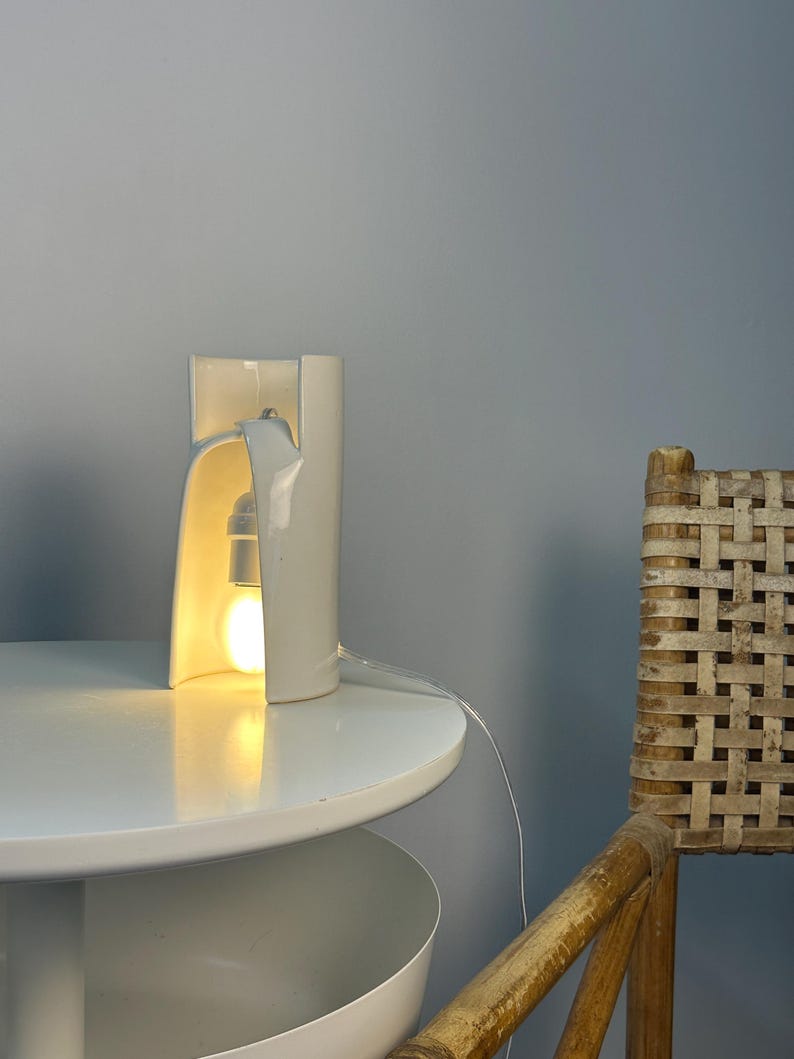 May include: A white ceramic table lamp with a curved, cylindrical structure that is open on one side. The lamp features a single light bulb emitting a warm glow from within the hollow center, connected by a thin, clear electrical cord.