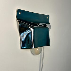 May include: A wall-mounted lamp featuring a deep teal, reflective, curved metal shade with a smooth, glossy finish. The fixture is secured with two visible silver screws at the top and includes a single round, frosted light bulb extending from the bottom.