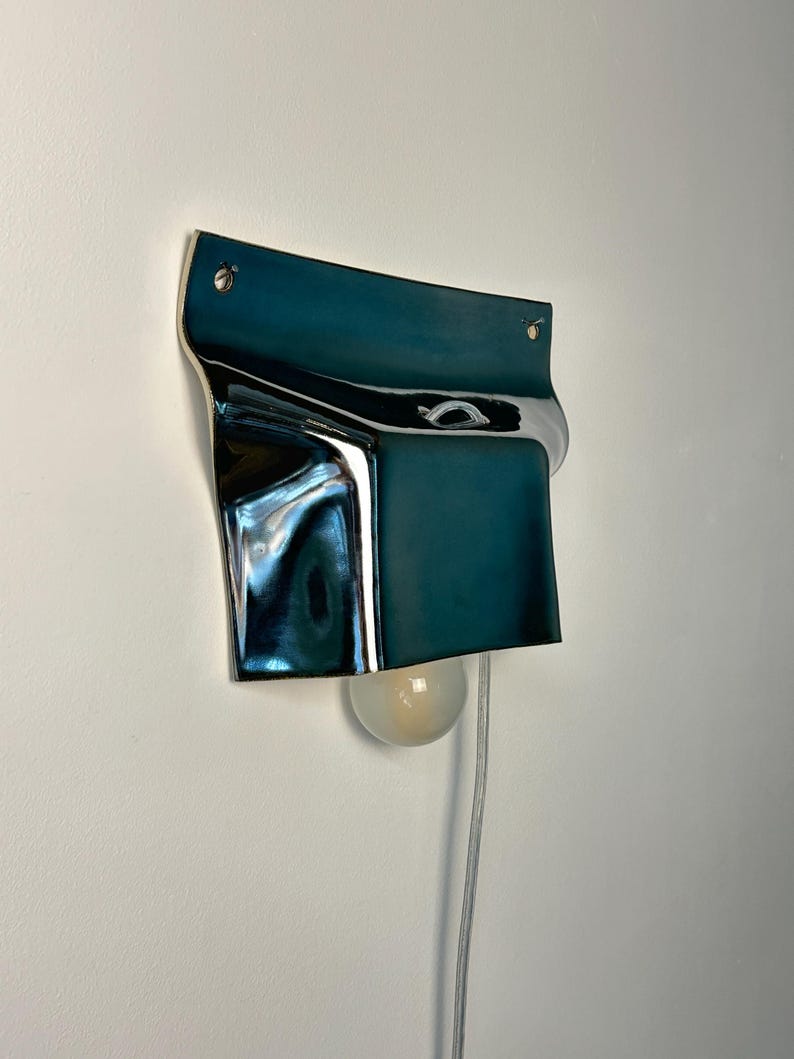 May include: A wall-mounted lamp featuring a deep teal, reflective, curved metal shade with a smooth, glossy finish. The fixture is secured with two visible silver screws at the top and includes a single round, frosted light bulb extending from the bottom.