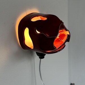 May include: A dark red wall sconce with an unusual, organic form. The sconce has several openings that allow light to shine through, creating a warm glow. The interior is a bright orange colour. A black base and a silver cord are attached.