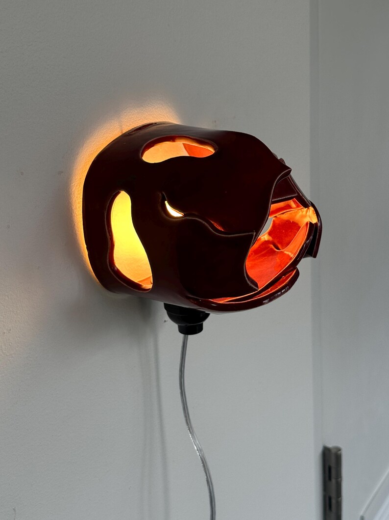May include: A dark red wall sconce with an unusual, organic form. The sconce has several openings that allow light to shine through, creating a warm glow. The interior is a bright orange colour. A black base and a silver cord are attached.