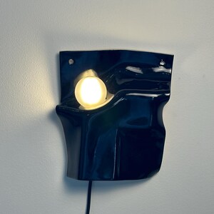 Puede incluir: Un aplique de pared con una pantalla de cer&aacute;mica azul marino brillante de forma irregular, plegada y org&aacute;nica. Una bombilla blanca redonda y brillante est&aacute; empotrada en la secci&oacute;n superior izquierda de la forma de cer&aacute;mica, con un cable negro.