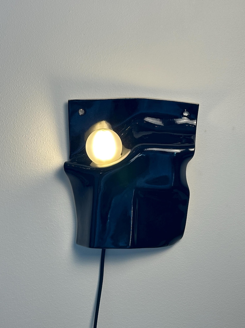 Puede incluir: Un aplique de pared con una pantalla de cer&aacute;mica azul marino brillante de forma irregular, plegada y org&aacute;nica. Una bombilla blanca redonda y brillante est&aacute; empotrada en la secci&oacute;n superior izquierda de la forma de cer&aacute;mica, con un cable negro.