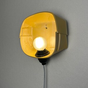 May include: A wall-mounted lamp featuring a curved, yellow-toned ceramic shade with a smooth interior finish. A single, round, frosted white light bulb is centered within the shade, attached to a black socket base with a thin, light-colored cord.
