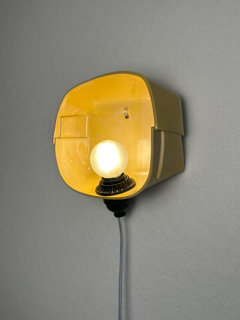May include: A wall-mounted lamp featuring a curved, yellow-toned ceramic shade with a smooth interior finish. A single, round, frosted white light bulb is centered within the shade, attached to a black socket base with a thin, light-colored cord.