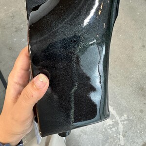 May include: A rectangular ceramic vessel with a glossy, dark black glaze that features subtle iridescent green and blue undertones. The surface is smooth and reflective, with slightly curved edges and a deep, dark finish that catches light.