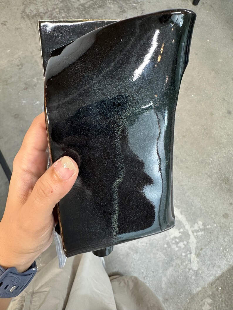 May include: A rectangular ceramic vessel with a glossy, dark black glaze that features subtle iridescent green and blue undertones. The surface is smooth and reflective, with slightly curved edges and a deep, dark finish that catches light.
