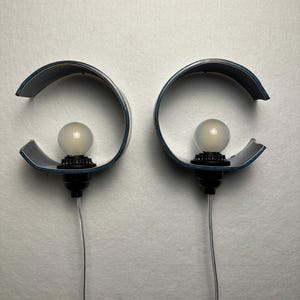 May include: A pair of wall-mounted lamps featuring curved, dark blue semi-circular shades. Each lamp has a black socket holding a round, frosted white light bulb. A thin, clear electrical cord extends downward from the base of each lamp fixture.