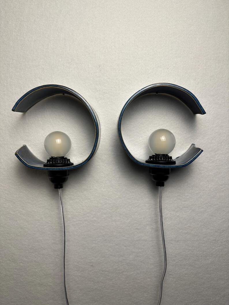 May include: A pair of wall-mounted lamps featuring curved, dark blue semi-circular shades. Each lamp has a black socket holding a round, frosted white light bulb. A thin, clear electrical cord extends downward from the base of each lamp fixture.