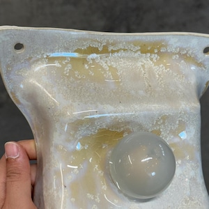 May include: A ceramic wall-mounted fixture with a glossy, cream-colored glaze featuring a textured, crystalline crackle pattern. The piece has a rectangular shape with rounded corners, two circular mounting holes, and a smooth, translucent orb.