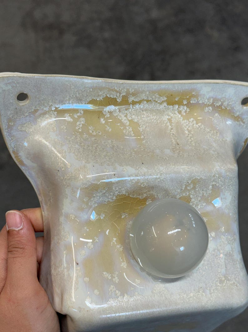 May include: A ceramic wall-mounted fixture with a glossy, cream-colored glaze featuring a textured, crystalline crackle pattern. The piece has a rectangular shape with rounded corners, two circular mounting holes, and a smooth, translucent orb.
