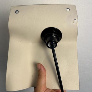 May include: A rectangular off-white textured panel featuring two circular metal grommets at the top corners. A black circular light socket fixture is mounted in the center, with a black electrical cord extending downward from the base.