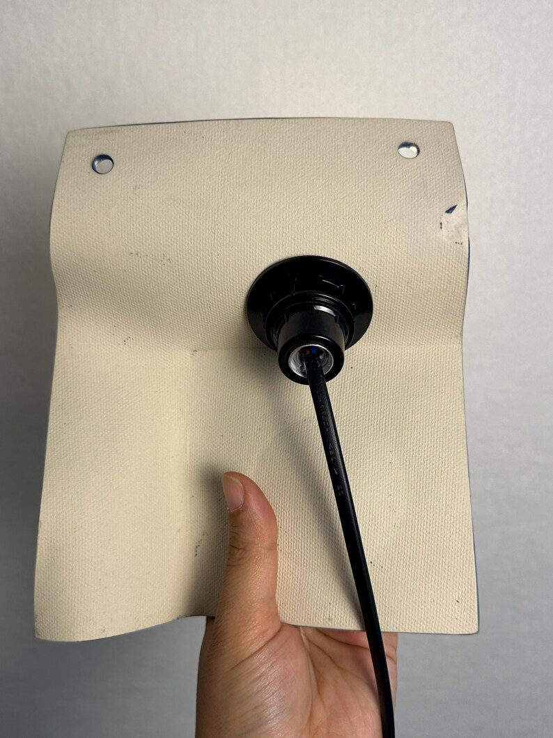 May include: A rectangular off-white textured panel featuring two circular metal grommets at the top corners. A black circular light socket fixture is mounted in the center, with a black electrical cord extending downward from the base.
