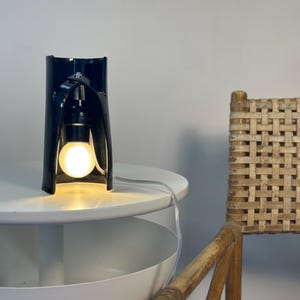 May include: A black ceramic table lamp with a curved, cylindrical structure that features an open front. The lamp houses a single glowing light bulb suspended from the top interior, connected by a clear electrical cord extending outward.