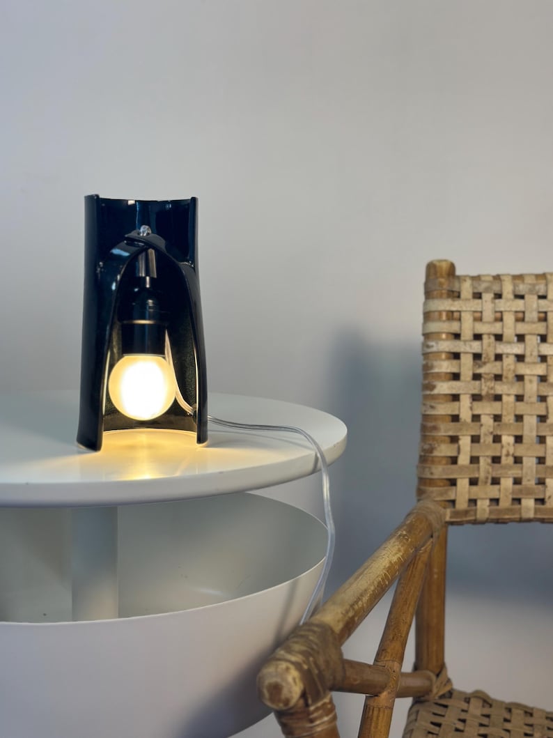 May include: A black ceramic table lamp with a curved, cylindrical structure that features an open front. The lamp houses a single glowing light bulb suspended from the top interior, connected by a clear electrical cord extending outward.