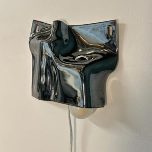 May include: A wall-mounted lamp featuring a reflective, metallic silver surface with an irregular, fluid, and undulating shape. The fixture includes a single light bulb extending from the bottom and a thin, light-colored power cord hanging down.