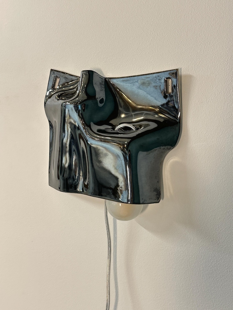 May include: A wall-mounted lamp featuring a reflective, metallic silver surface with an irregular, fluid, and undulating shape. The fixture includes a single light bulb extending from the bottom and a thin, light-colored power cord hanging down.