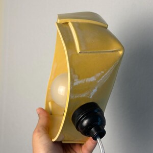 May include: A yellow ceramic wall lamp with a curved, rectangular form and a smooth, glossy finish. The fixture features a central white light bulb socket and a black plastic electrical component attached to the side with a clear power cord.